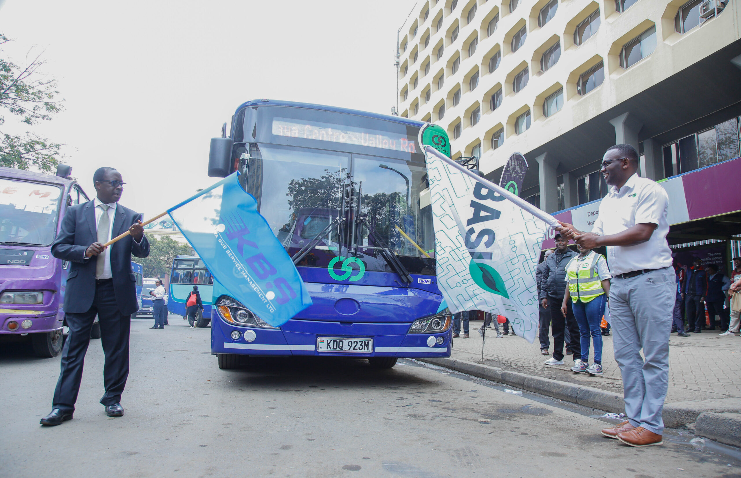 Officials waving KBSM flags beside a blue city bus, registration KDQ 923M, at a route launch ceremony in Nairobi CBD